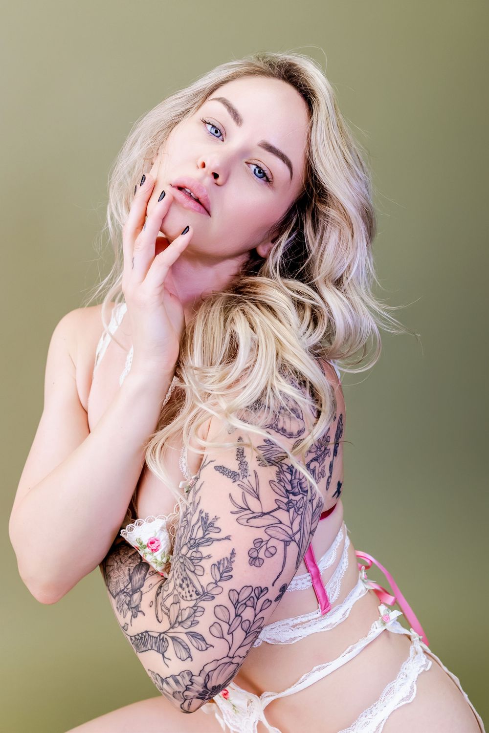 Boudoir photo of woman in white lingerie showing floral arm tattoo against olive green backdrop.