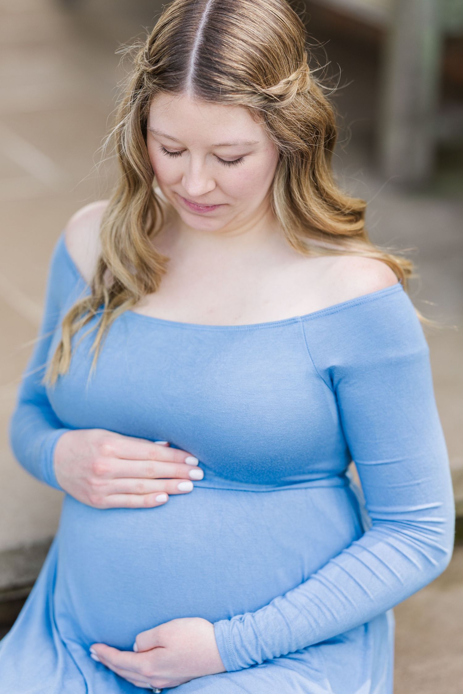 A pregnant woman in a light blue off-shoulder dress gently cradles her baby bump while looking down with a soft smile at Chicago Botanic Gar
