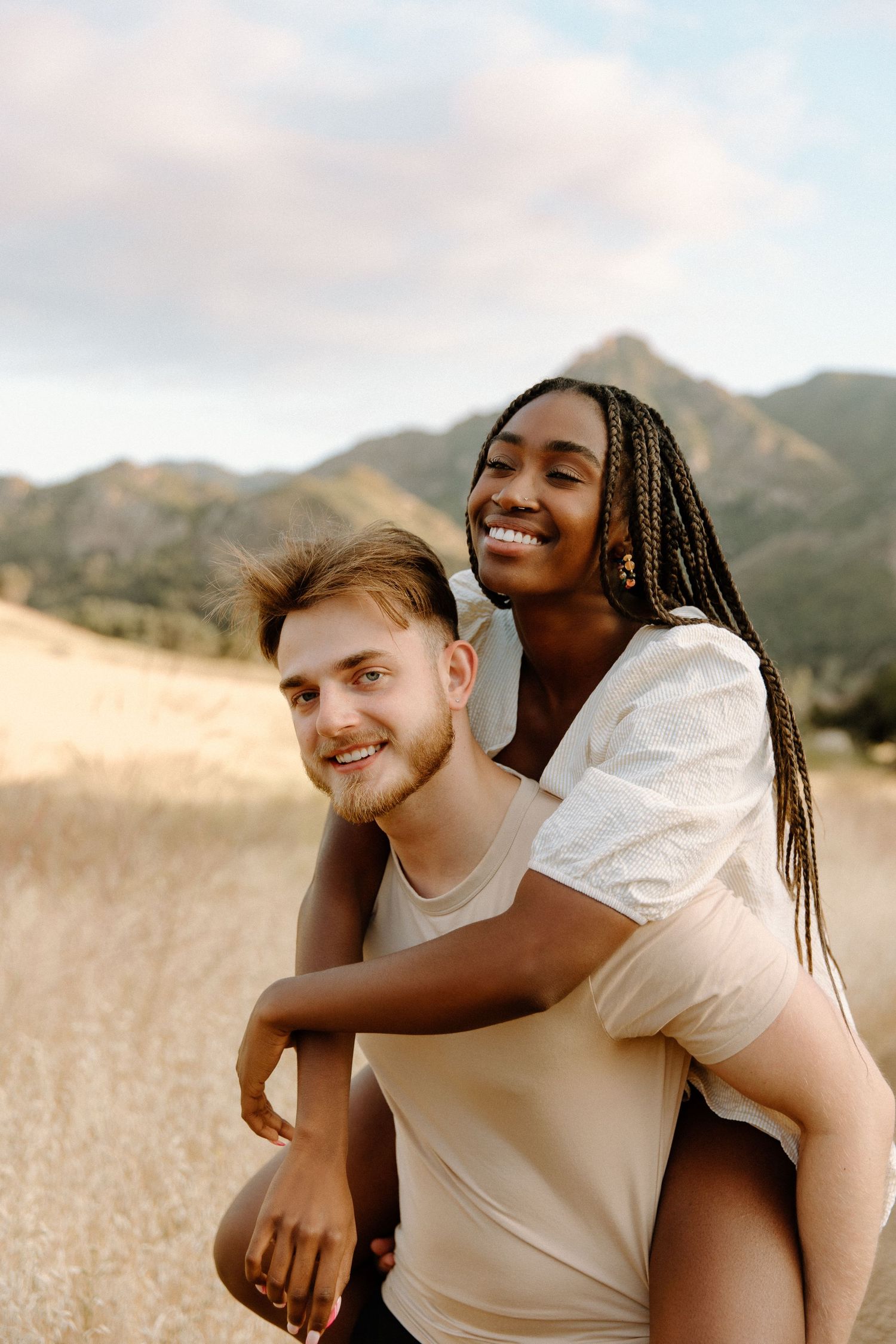 A happy couple shares a playful piggyback moment in a golden field with mountains in the background.