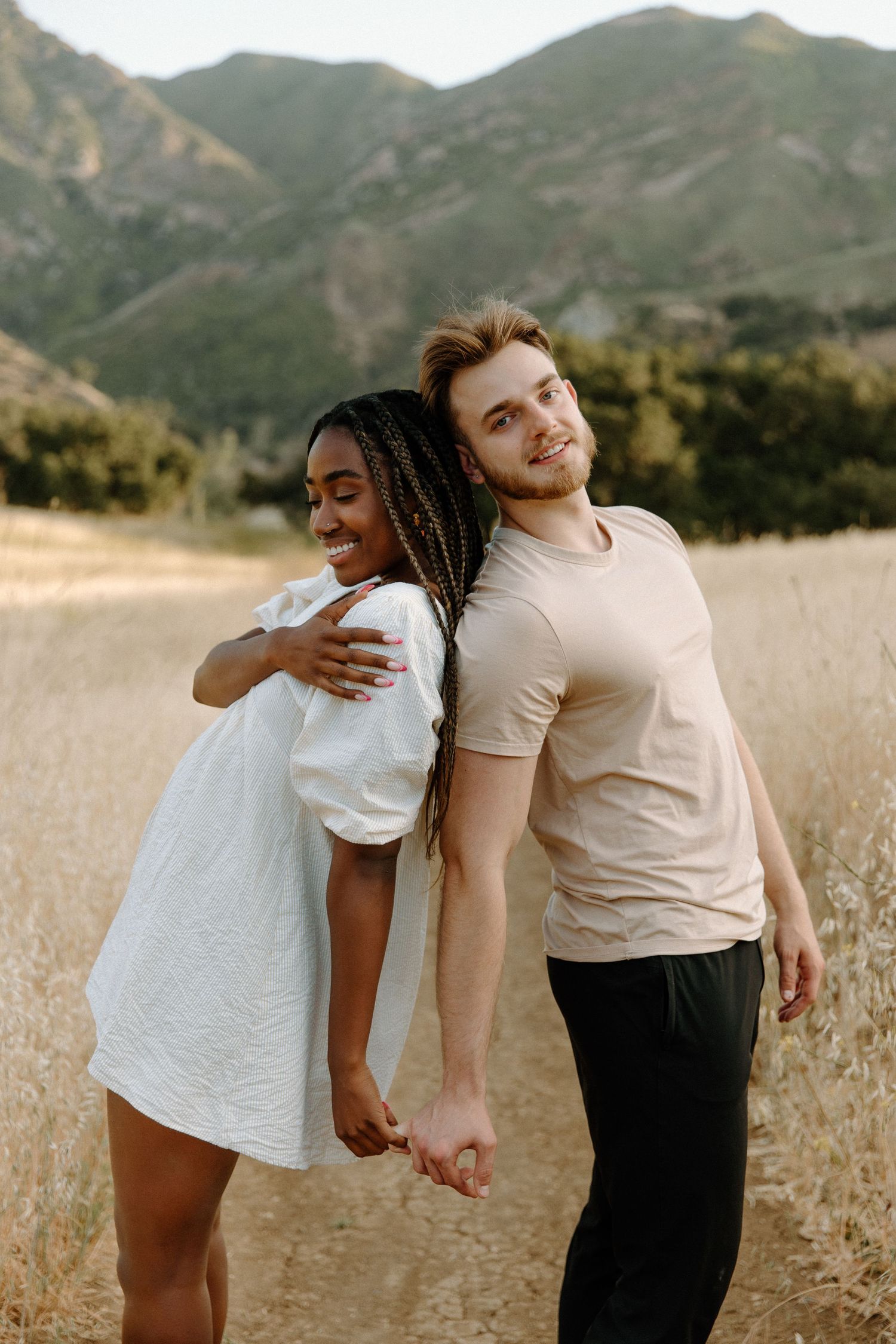 A happy couple poses back-to-back in a field with scenic mountains in the background during golden hour.