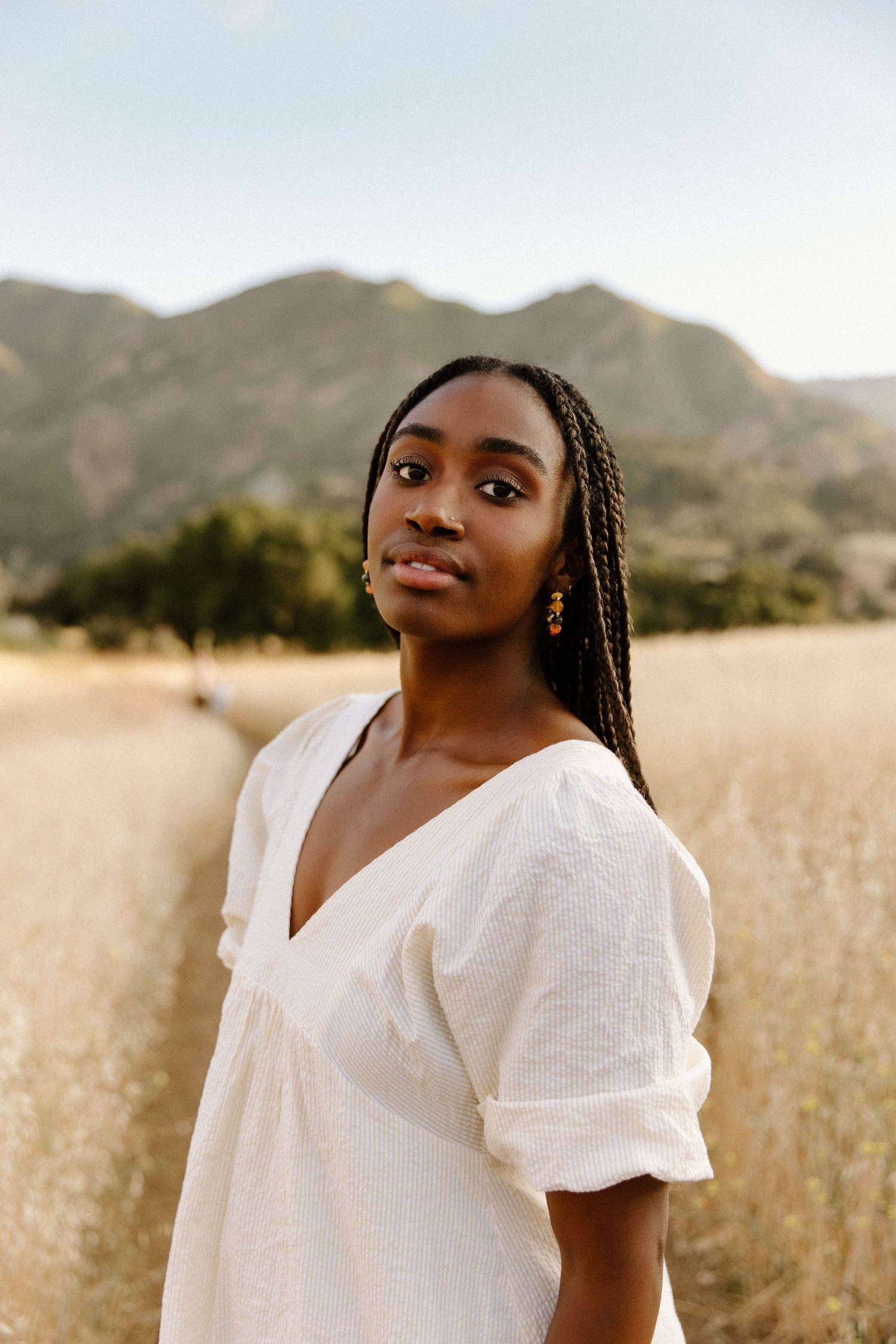 Person wearing a white dress poses in a golden field with mountains in the background during sunset.
