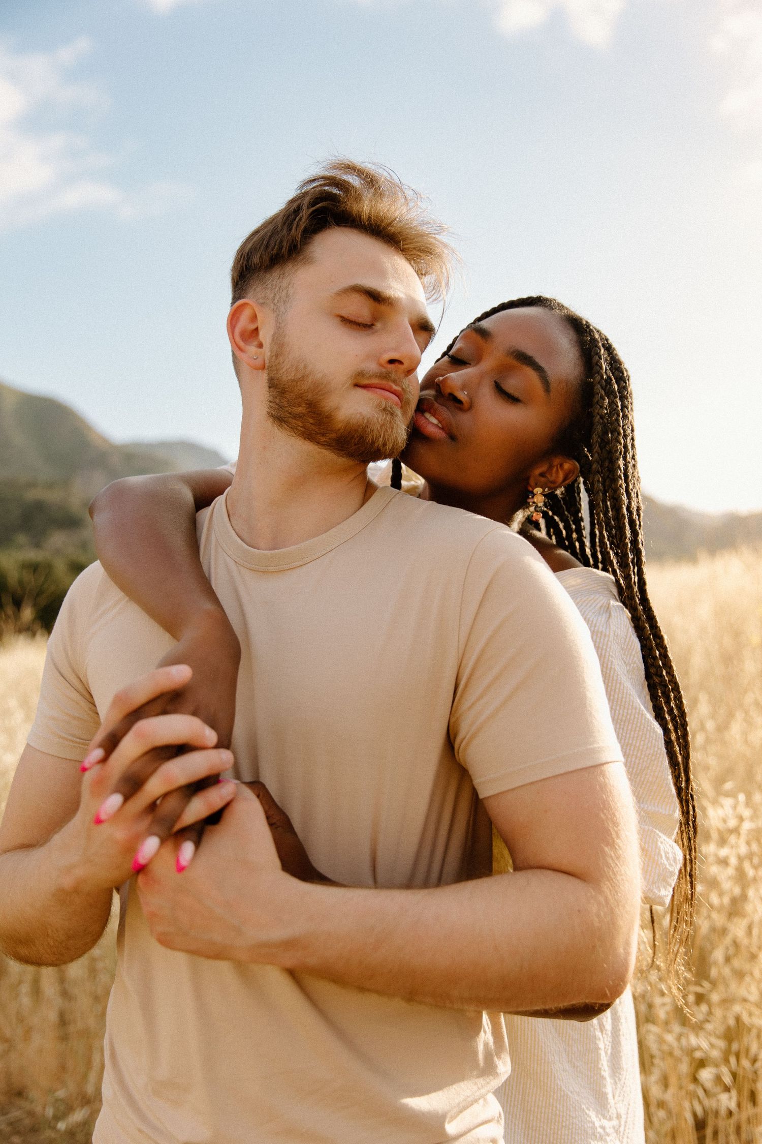 An interracial couple embraces tenderly in a sun-lit field of tall grass with mountains in the background.