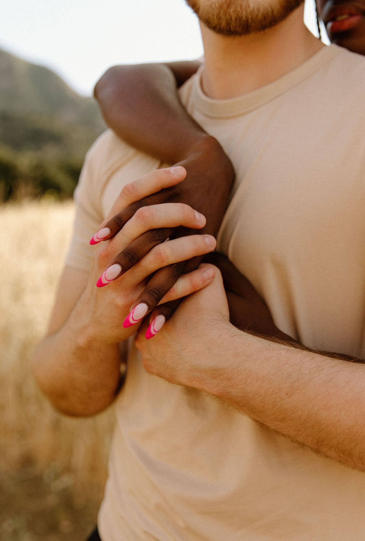 A close-up of intertwined hands with pink painted nails against a beige shirt and outdoor nature background.