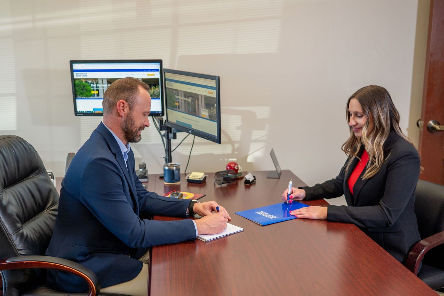 Two professionals having a discussion at desk with computer monitors in office setting.