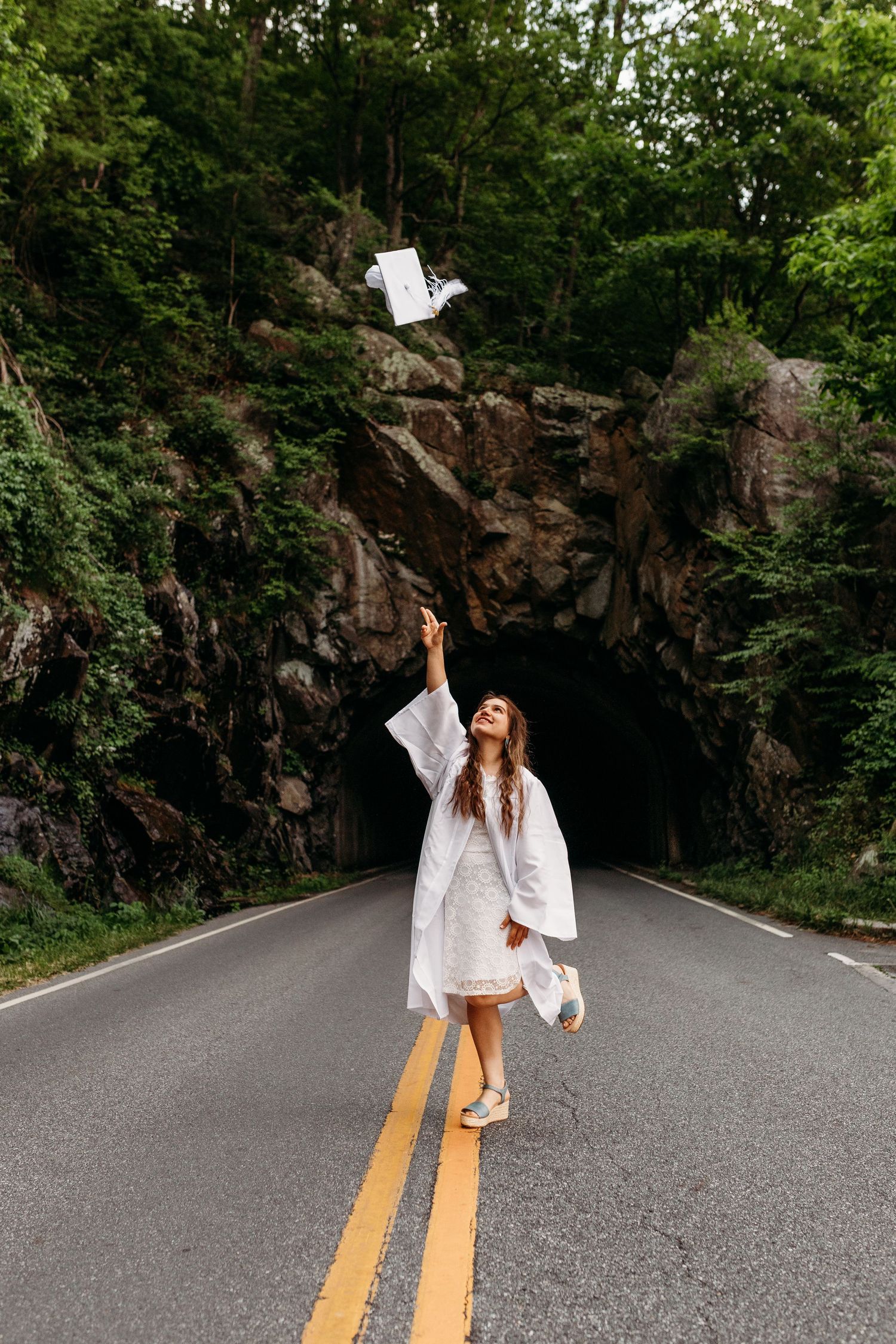 Graduate in white dress throws diploma papers in front of rock tunnel on rural road, celebrating achievement.