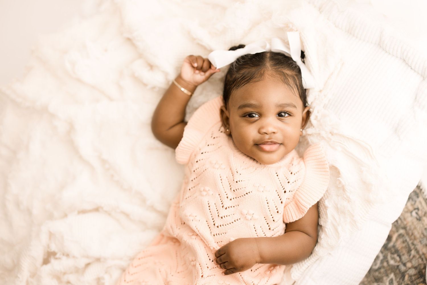 Baby wearing peach colored dress lies on soft white blanket smiling at camera.
