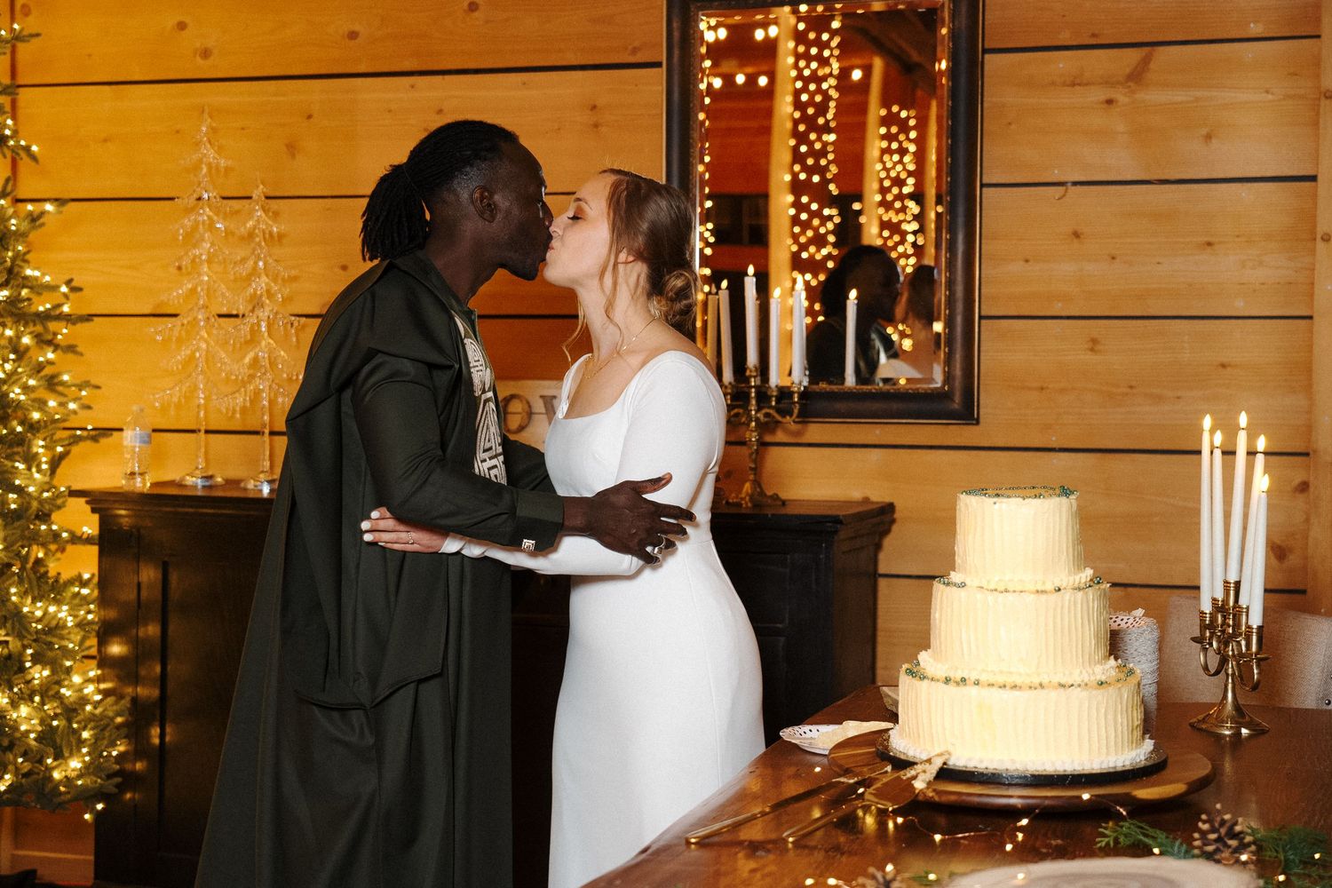 A romantic wedding scene with a three-tiered white cake, candelabra, and holiday decor in a rustic wooden cabin setting.