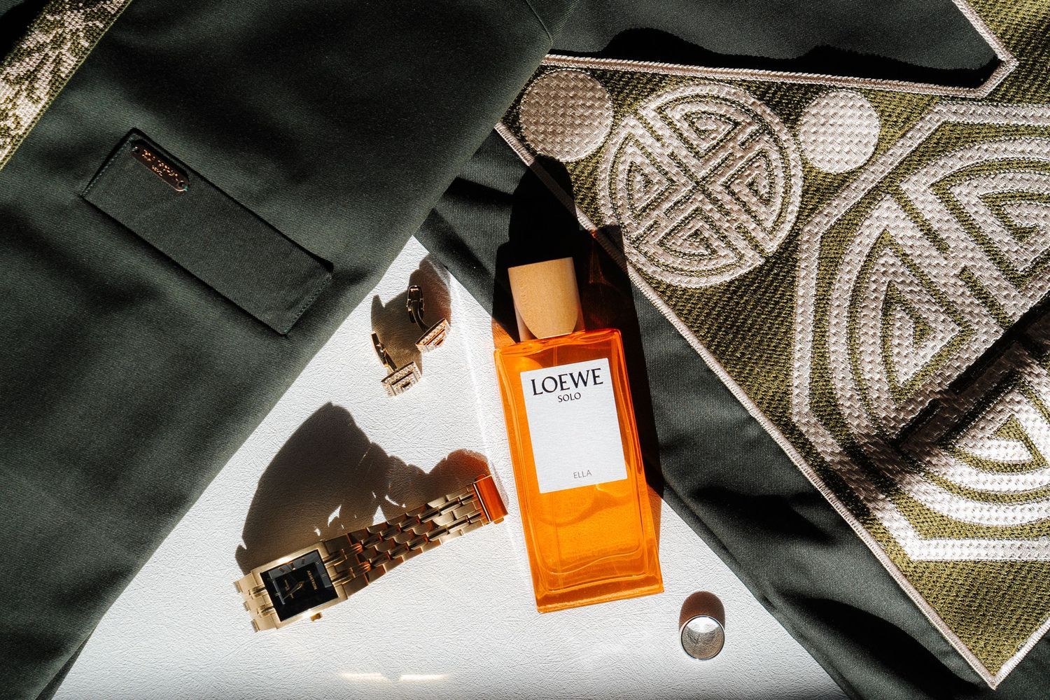 Flat lay of an orange fragrance bottle next to green fabric and geometric patterned textile with scattered chocolates.
