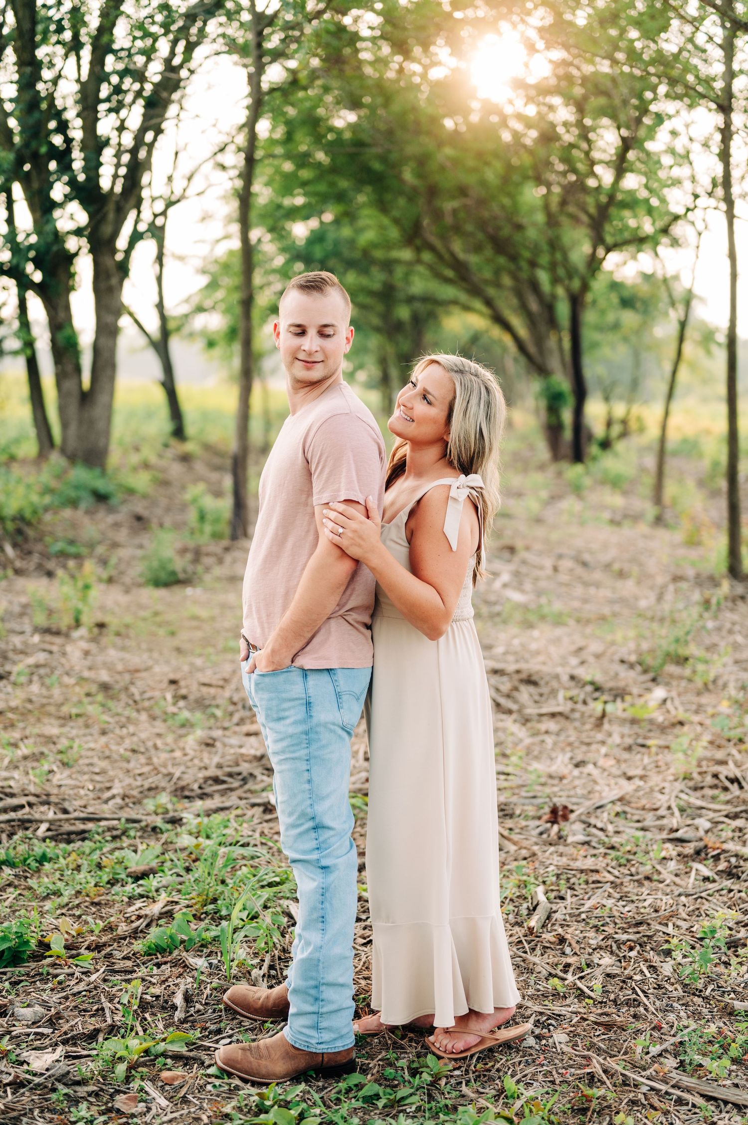 Romantic couple embraces while standing in a sunlit grassy field with green trees in the background.