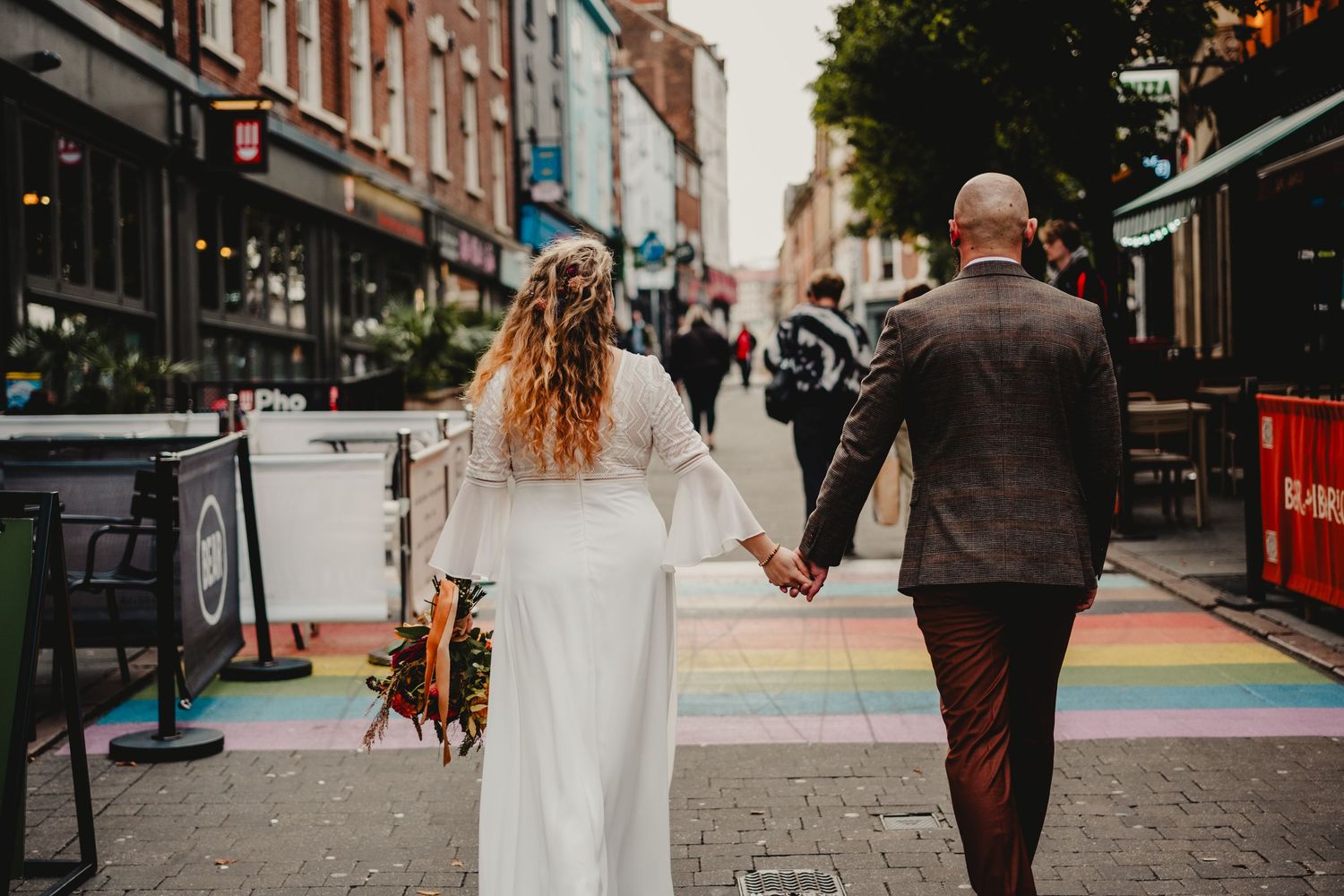 A couple walks hand in hand down a colorful rainbow-painted city street lined with shops and businesses.
