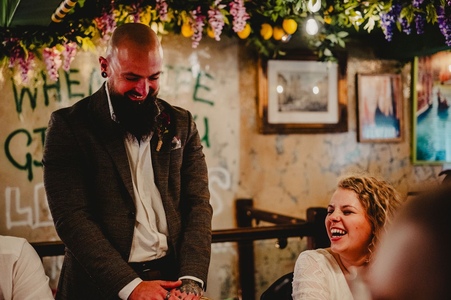 Two people share a joyful candid moment at what appears to be a rustic wedding venue decorated with floral arrangements.