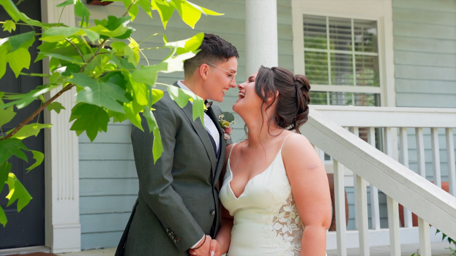 A romantic couple in wedding attire share a happy moment on a white porch near green foliage.