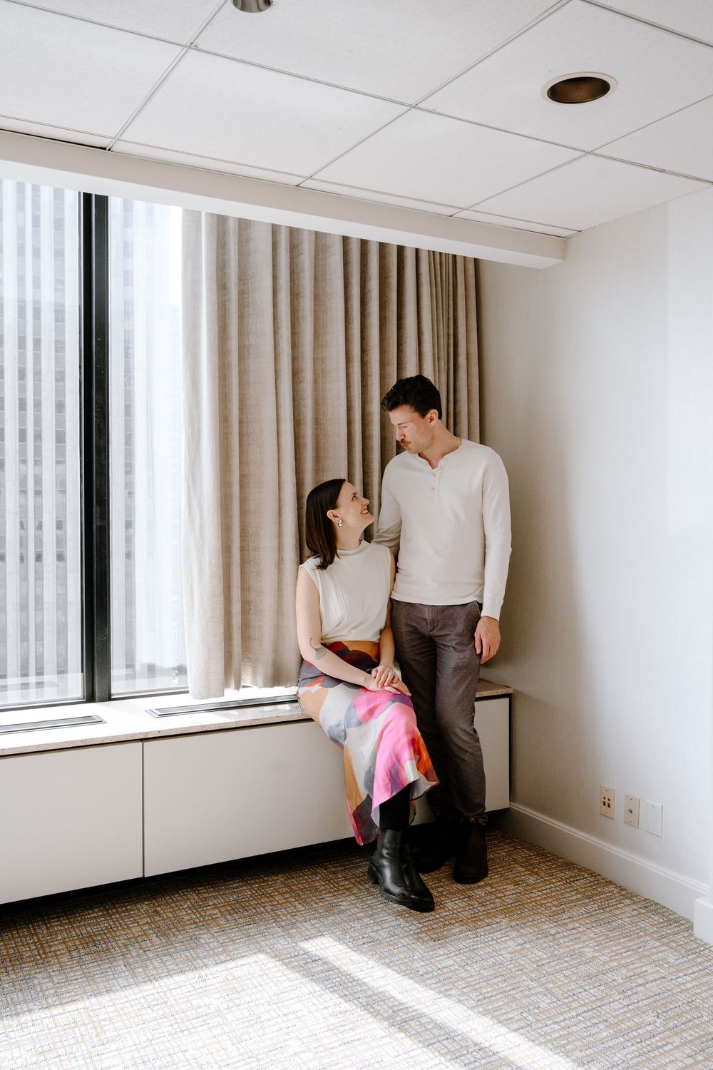 A couple shares an intimate moment near a window in a bright minimalist room with beige curtains and carpeted floors.