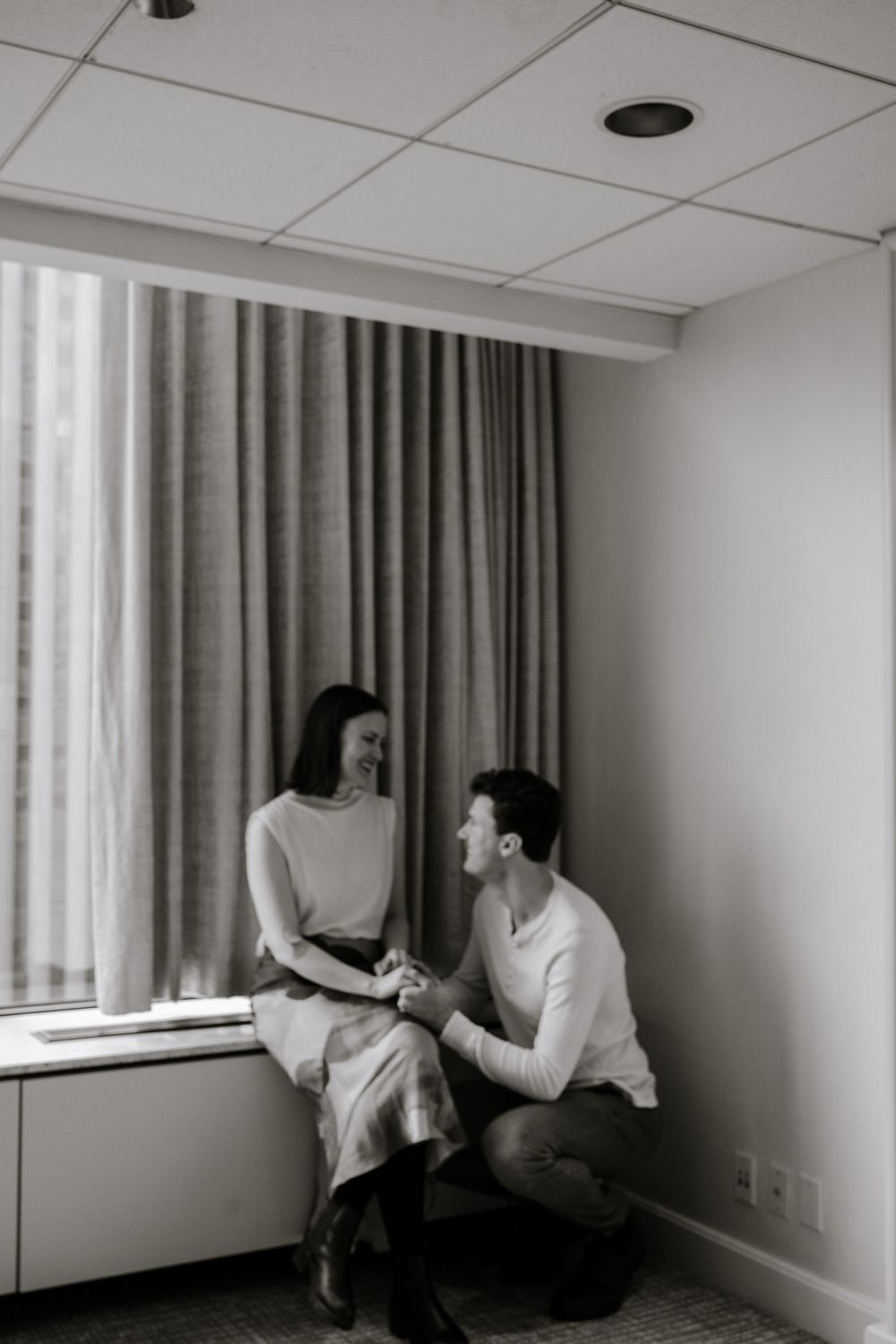 A black and white intimate moment between a couple near a window, with dramatic curtains and soft lighting.