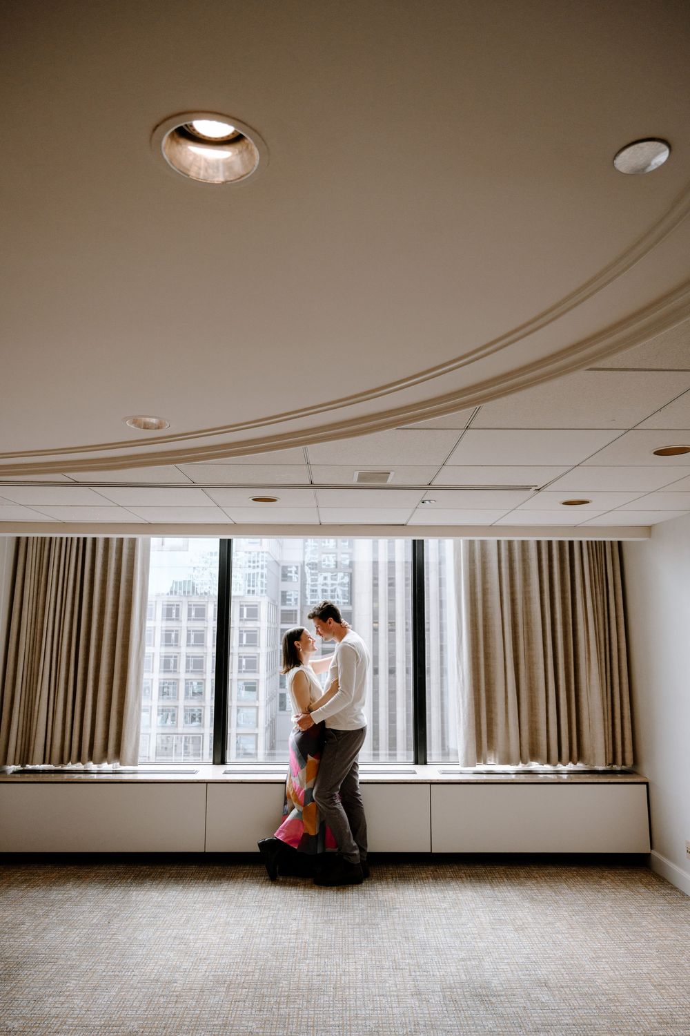 A couple embraces in silhouette by a large window in an empty room with modern architectural details and curtains.