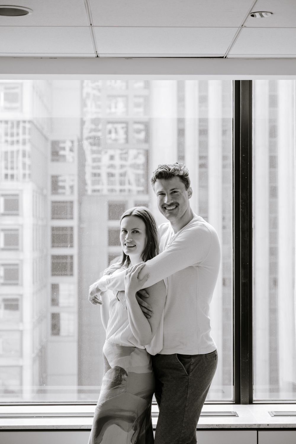 A black and white photo of a couple posing next to a large window with a city skyline view.