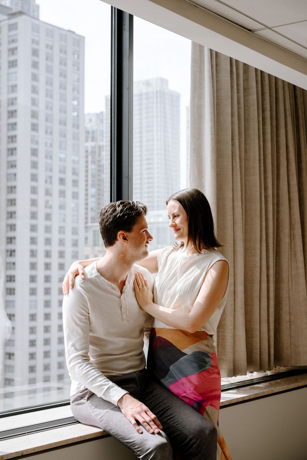 A couple shares an intimate moment while sitting by a large window overlooking city high-rises on a bright day.