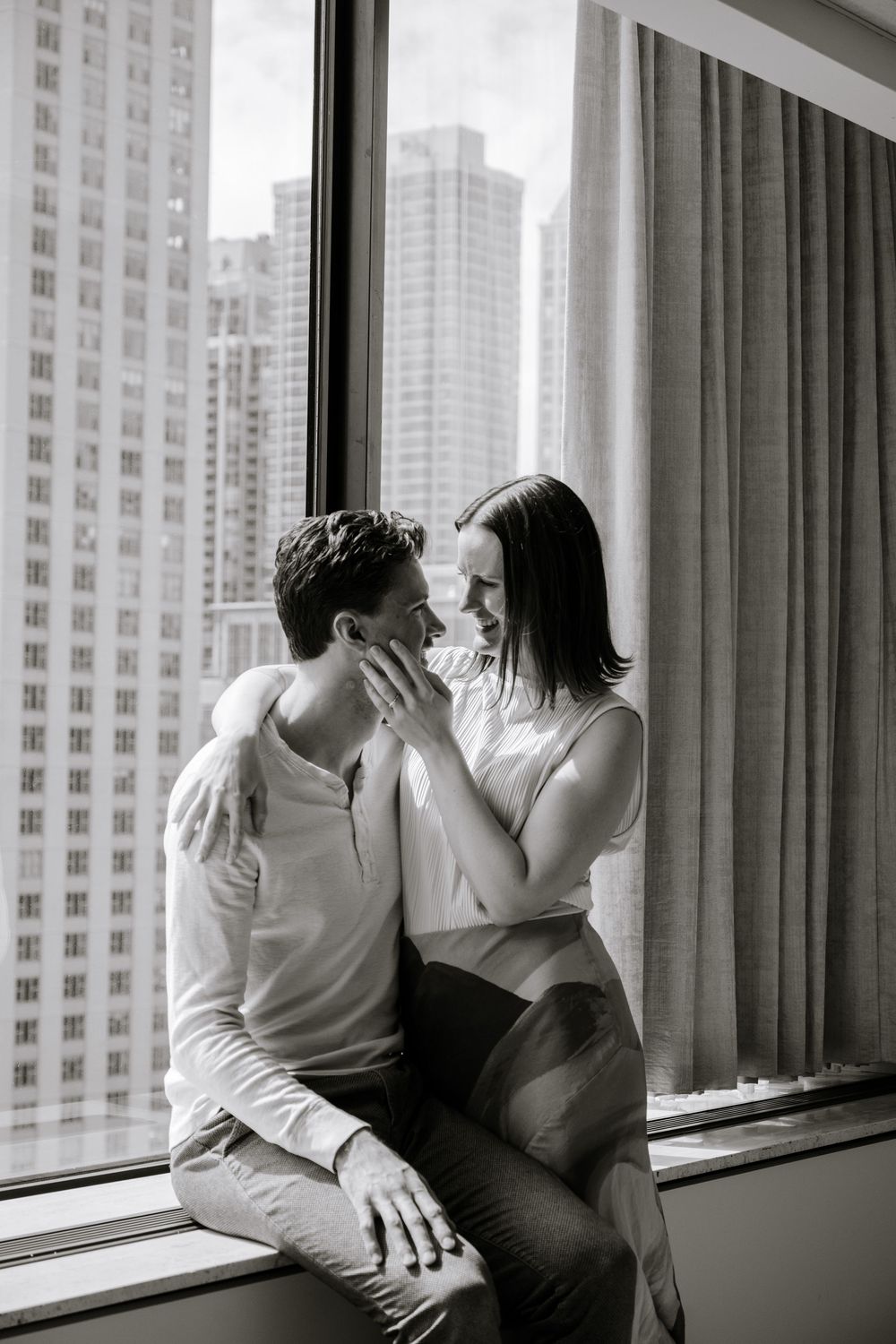 A romantic couple share an intimate moment by a window overlooking city high-rises in a black and white photograph.