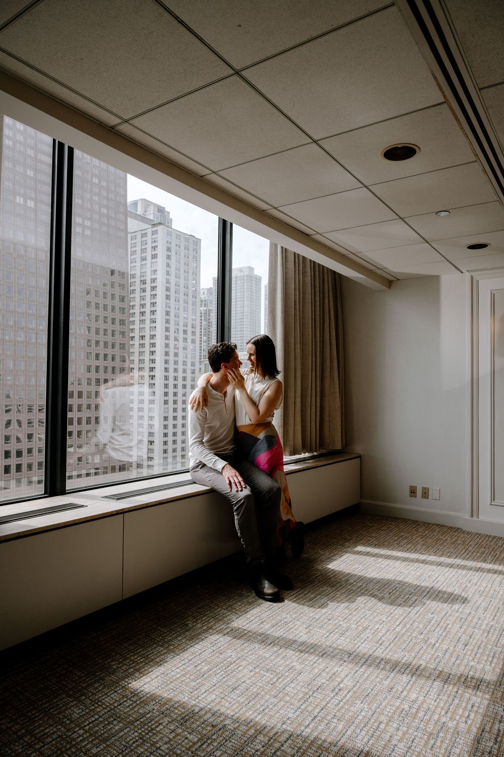 Two people share an intimate moment by a large window overlooking city buildings in a spacious modern room.