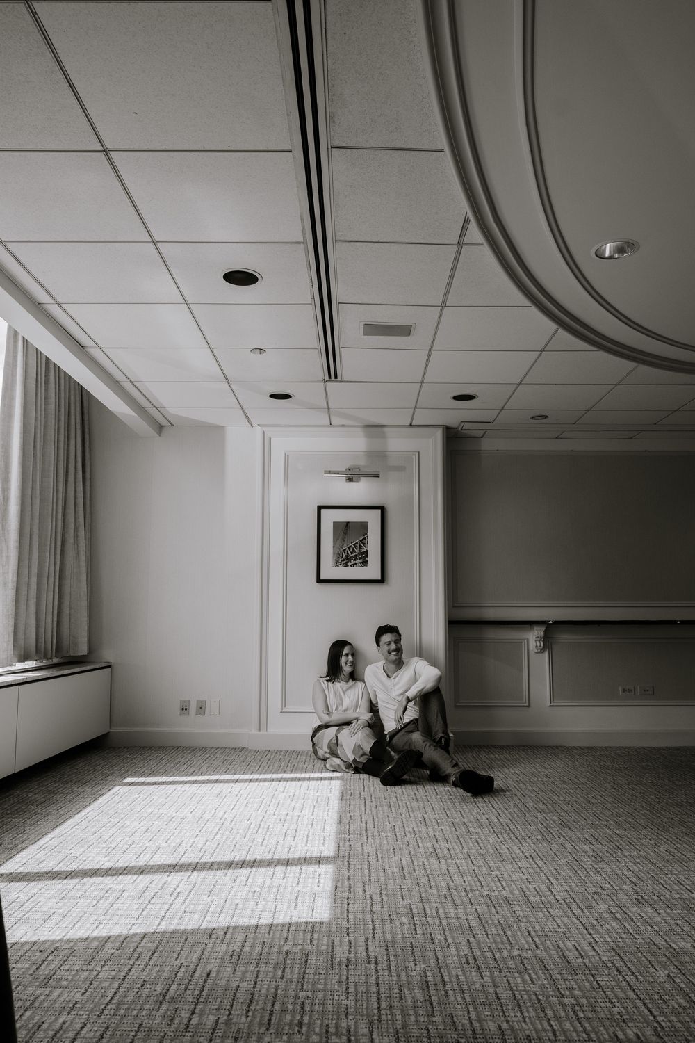 A black and white photo of a couple sitting on a carpeted floor in an empty room with a framed artwork on the wall.