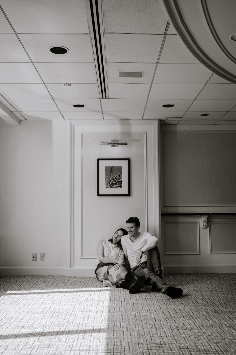 Black and white photograph showing a person sitting on the floor against a white wall with wainscoting in a minimalist interior.