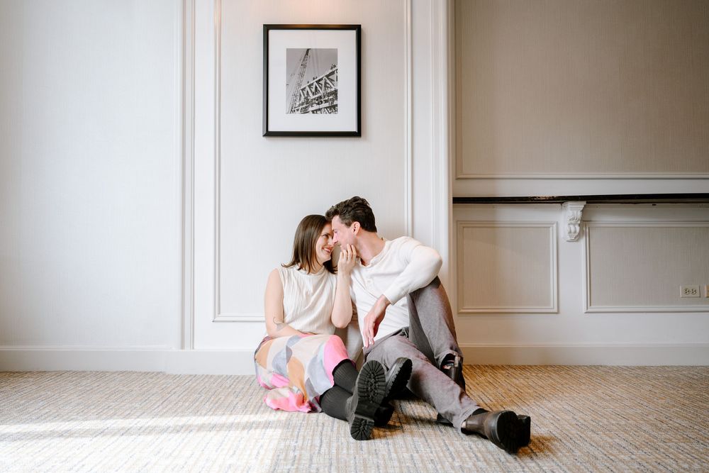 Affectionate couple sits together on hardwood floor against white wall with framed artwork.