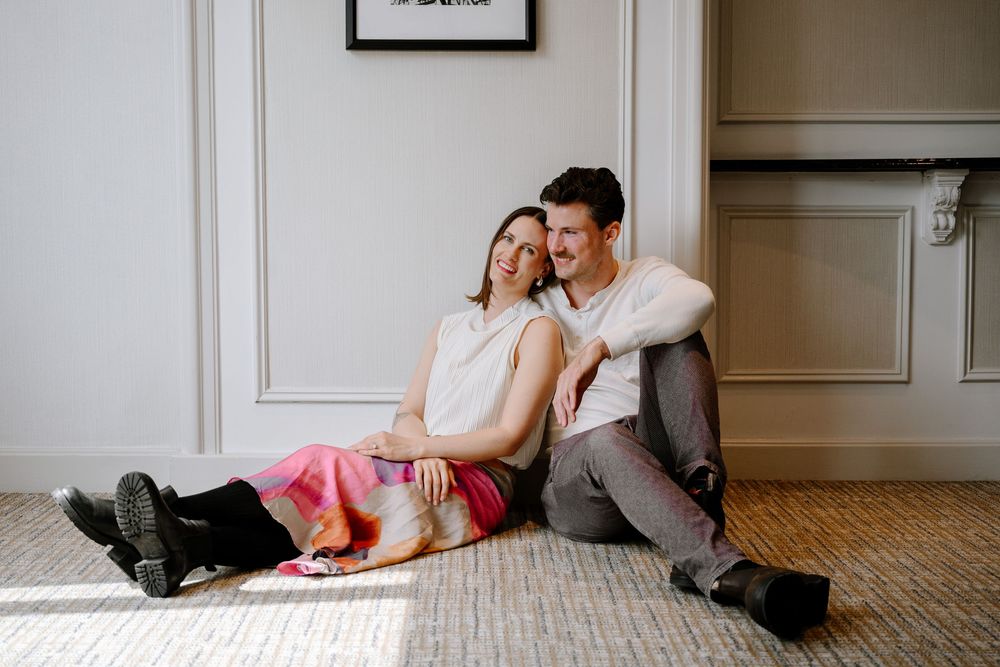 Couple sitting on floor against white wall, laughing and embracing in casual romantic moment.
