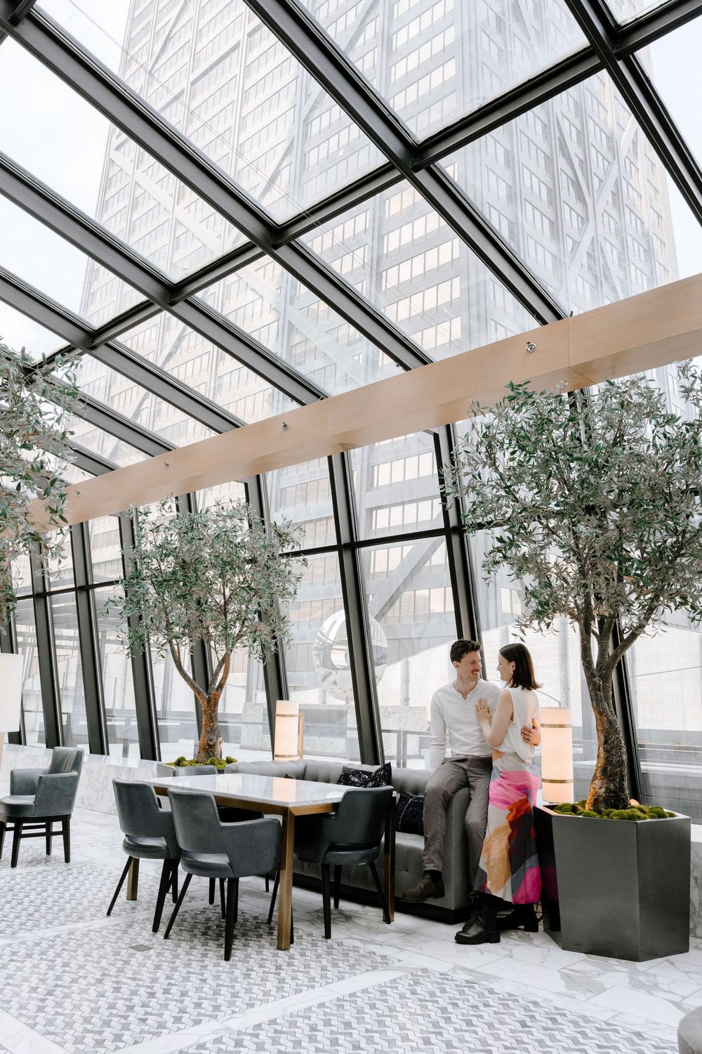 Modern restaurant interior with glass ceiling, indoor trees, and stylish furniture in a bright urban space.