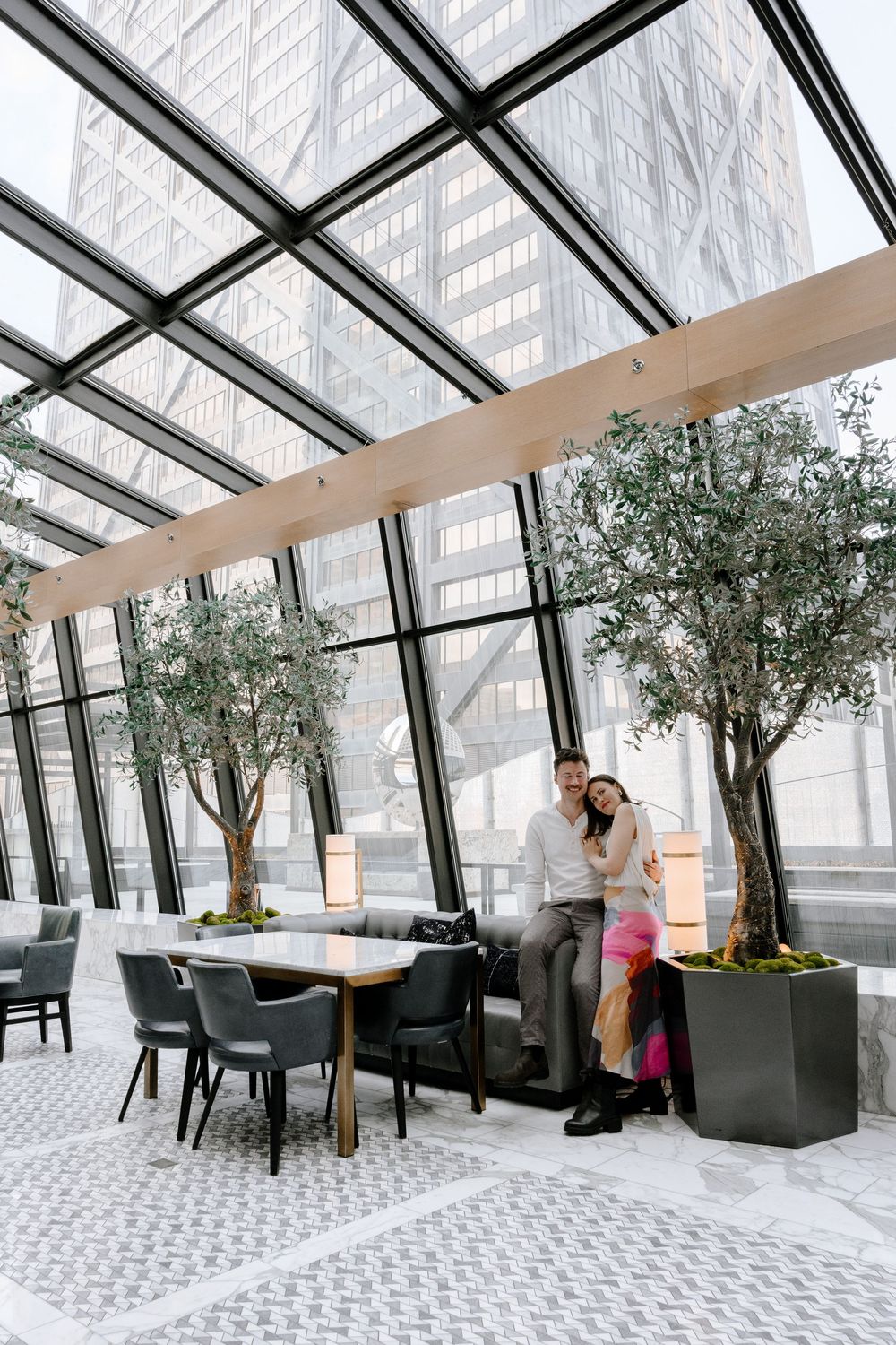 Modern restaurant interior with glass ceiling, potted olive trees, geometric floor tiles and contemporary dining furniture.