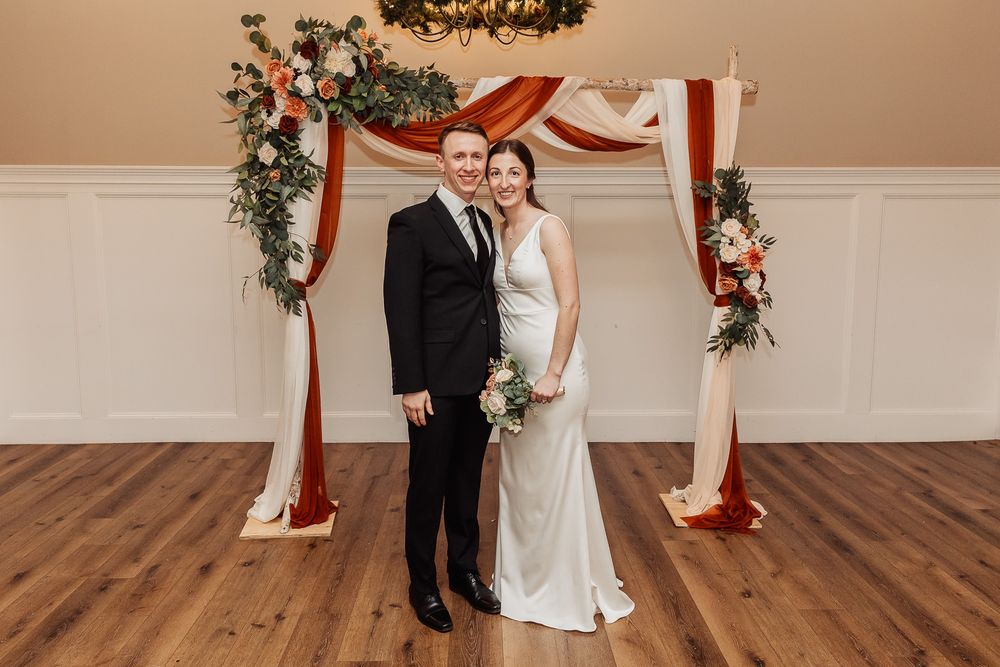 A couple poses beneath a red and white wedding arch adorned with greenery and flowers on a hardwood floor.