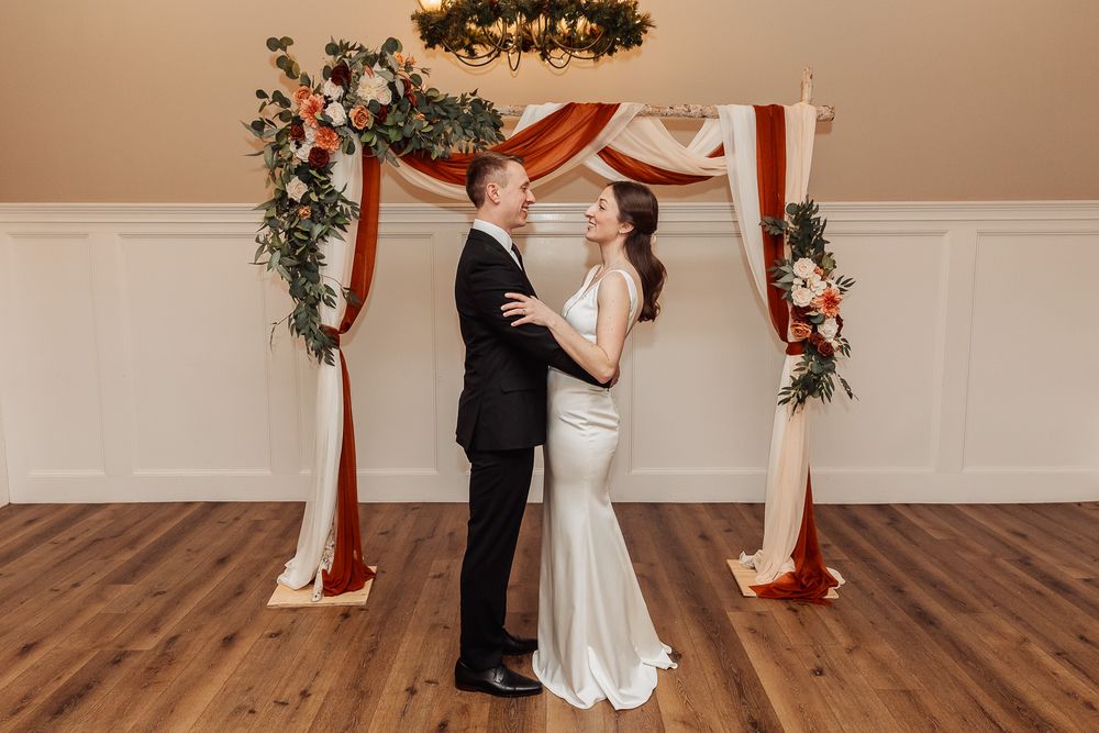 A couple stands under an elegant wooden arch decorated with red drapes and floral arrangements at an indoor venue.