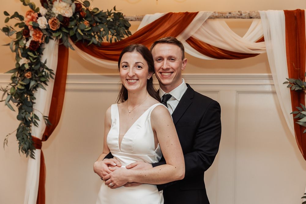 A couple embracing under an elegant wedding arch decorated with burgundy draping and greenery at an indoor venue.
