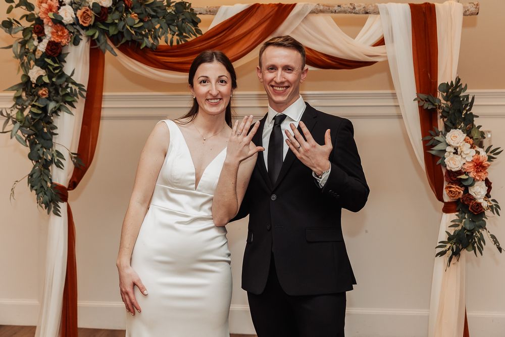 A couple shows off their rings at their wedding ceremony under an elegant arch decorated with coral and white flowers.
