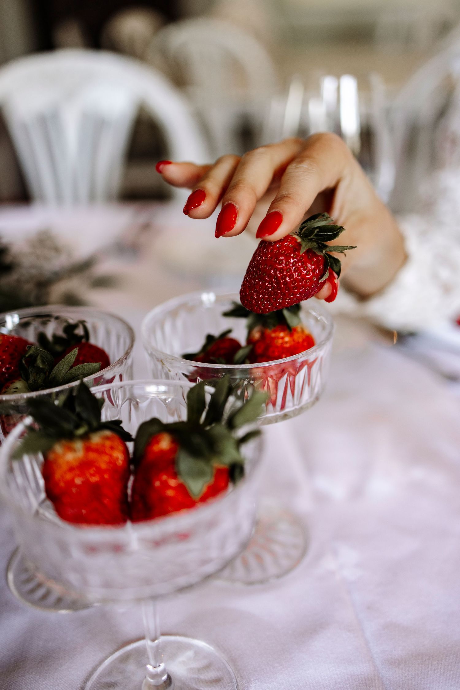 Fresh red strawberries in elegant crystal glasses with a hand reaching for one against a white tablecloth.