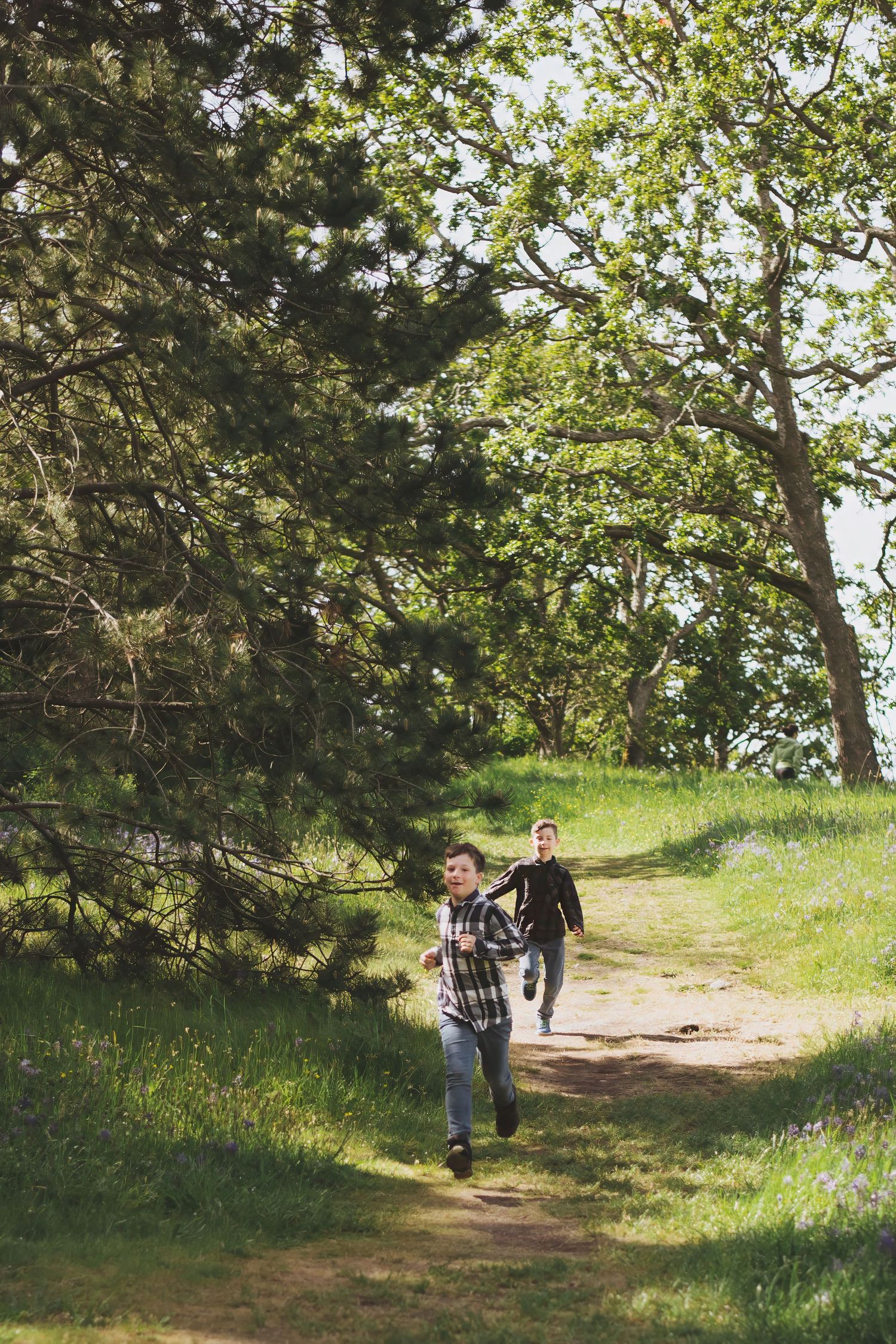 Two boys running along a sunny dirt trail through green trees and grass on a scenic nature path.
