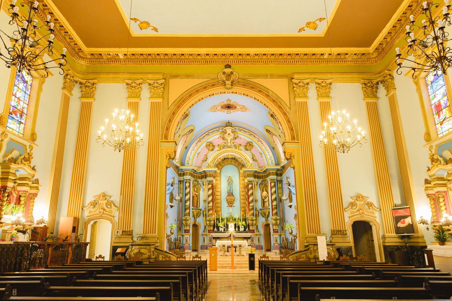 Interior view of an ornate yellow Catholic church with golden columns, crystal chandeliers and decorative altar in Hawaii.