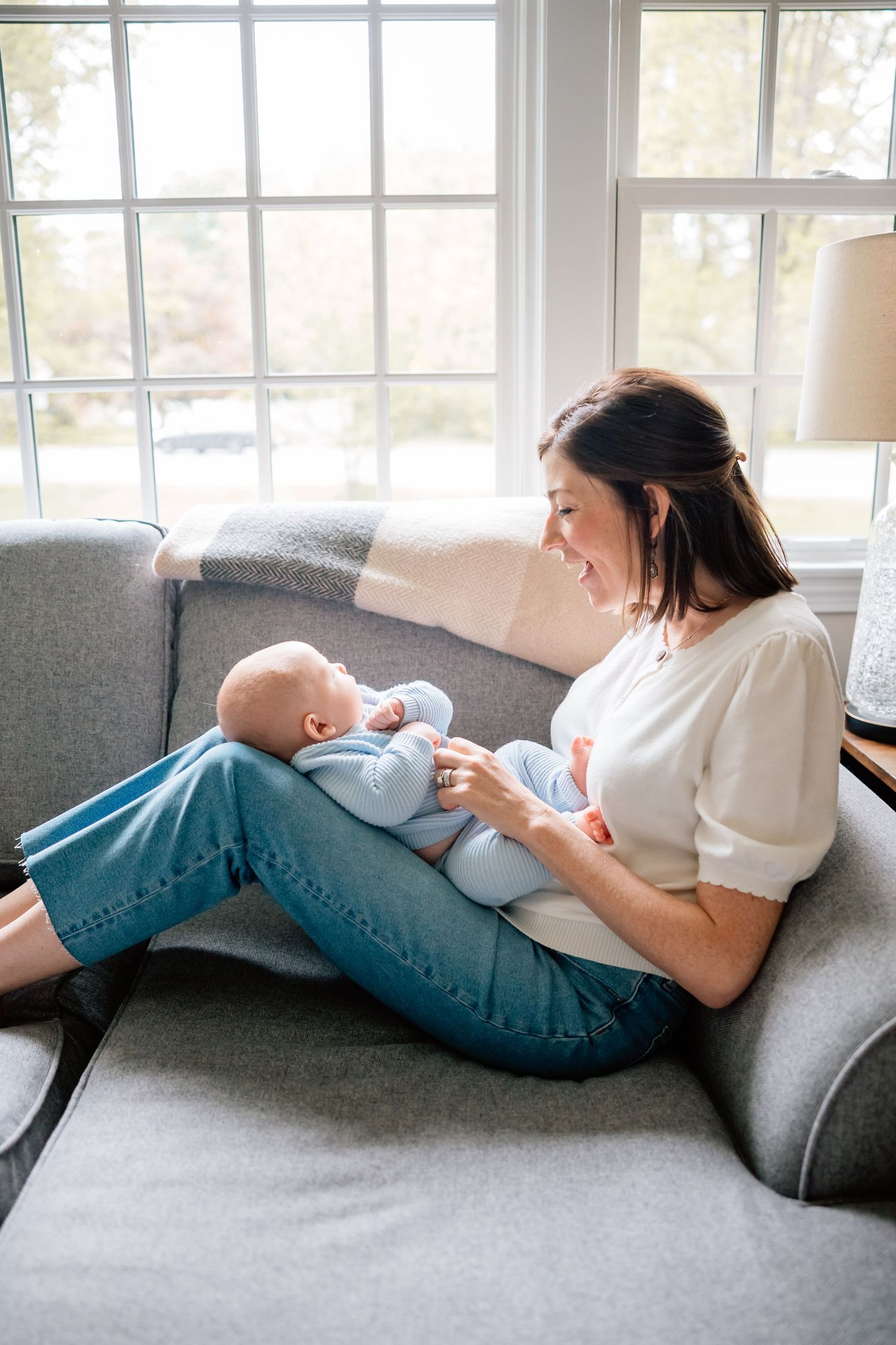 Tender moment on a gray couch by sunny windows as mother cuddles and feeds infant in blue sleepwear.