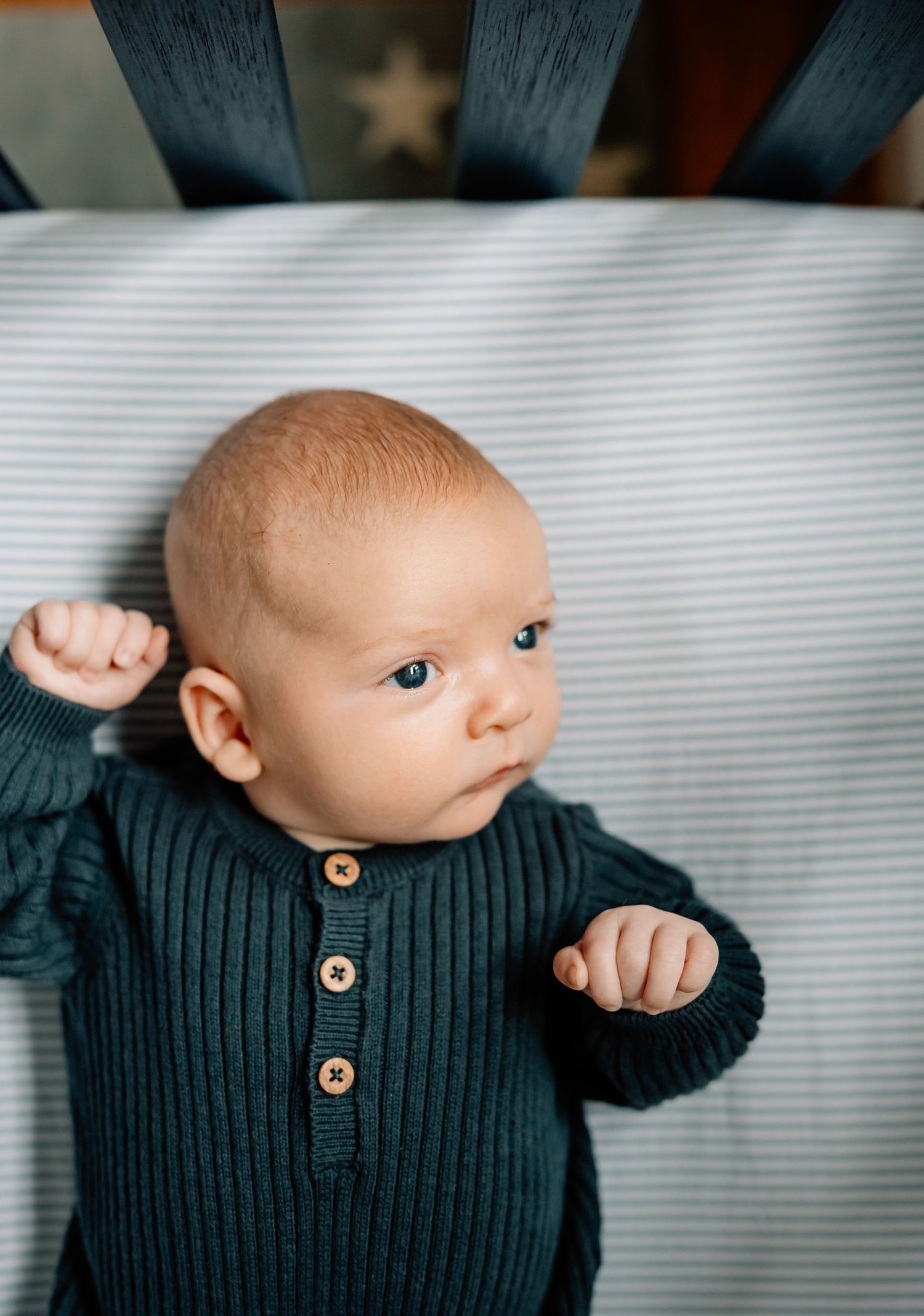 A baby in a dark green ribbed sweater with wooden buttons looks to the side while lying on striped bedding.