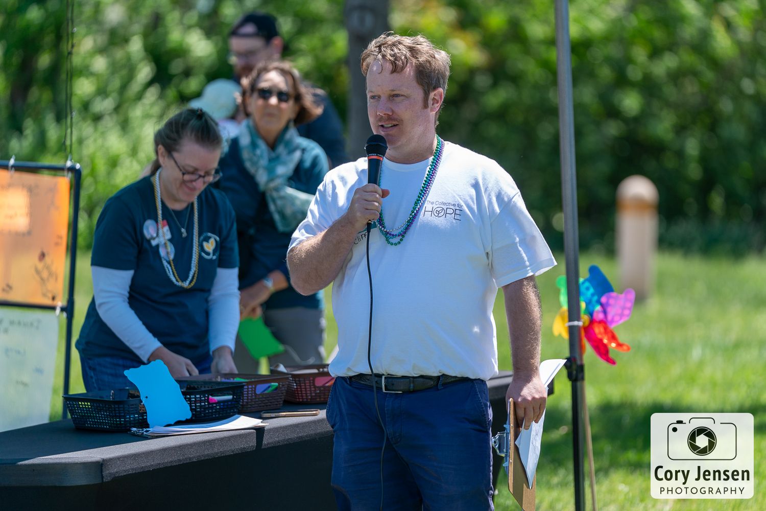 A person speaks into a microphone outdoors at a summer event with green trees and people in background.