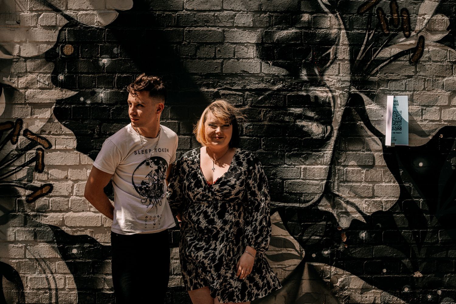 Two people pose in front of a dramatic black and white graffiti mural on a brick wall.