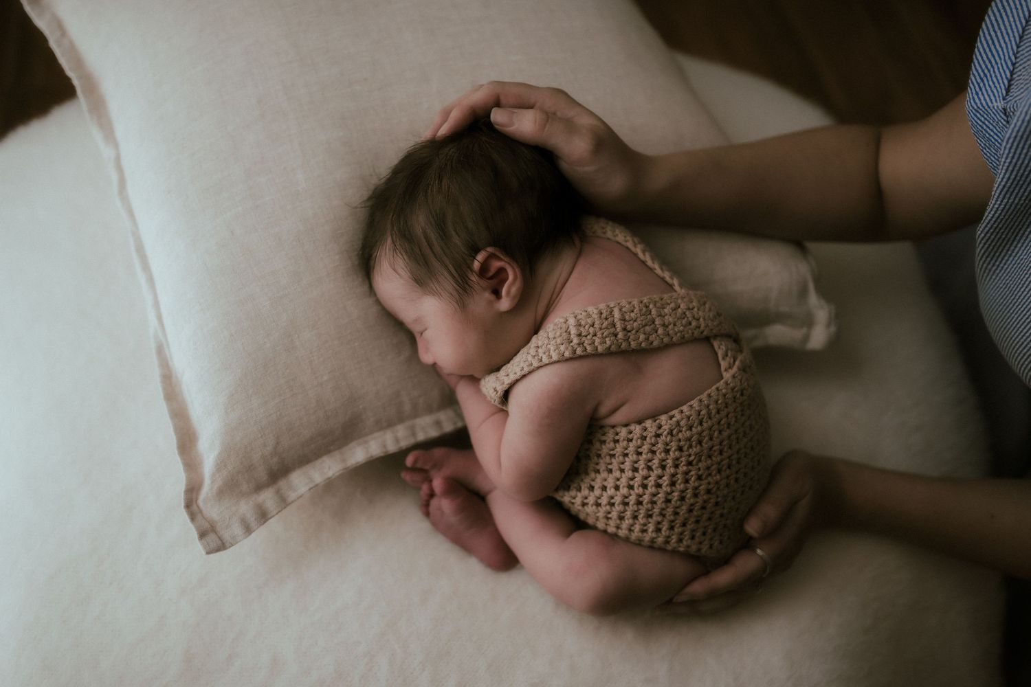 A sleeping baby wearing a knitted romper lies curled up on a beige cushion with a gentle hand touching their head.