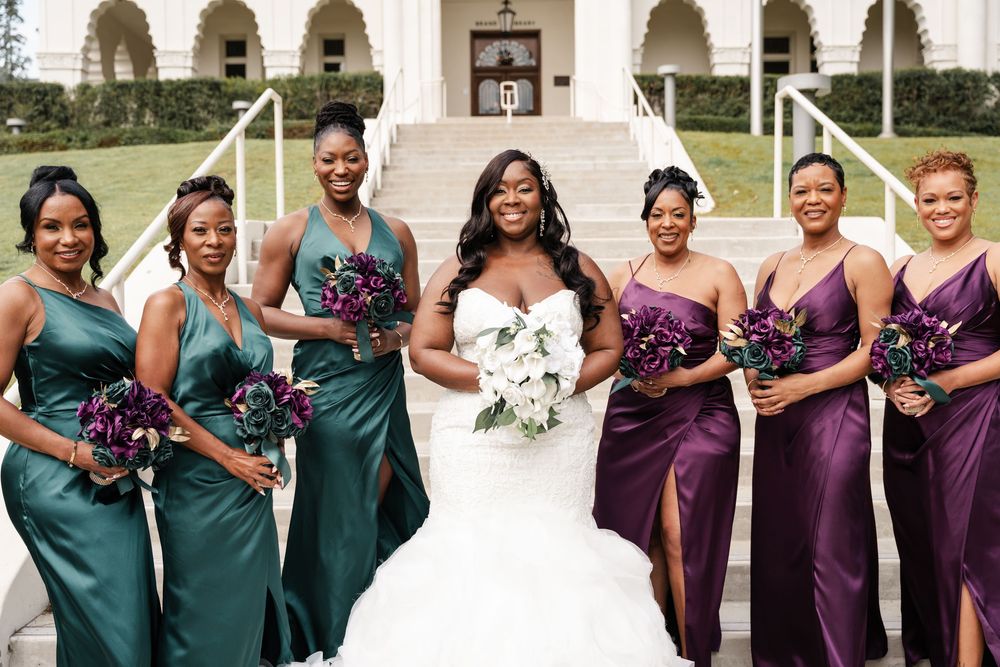 A bridal party wearing jewel-toned dresses in emerald and purple stand together on white stone steps.