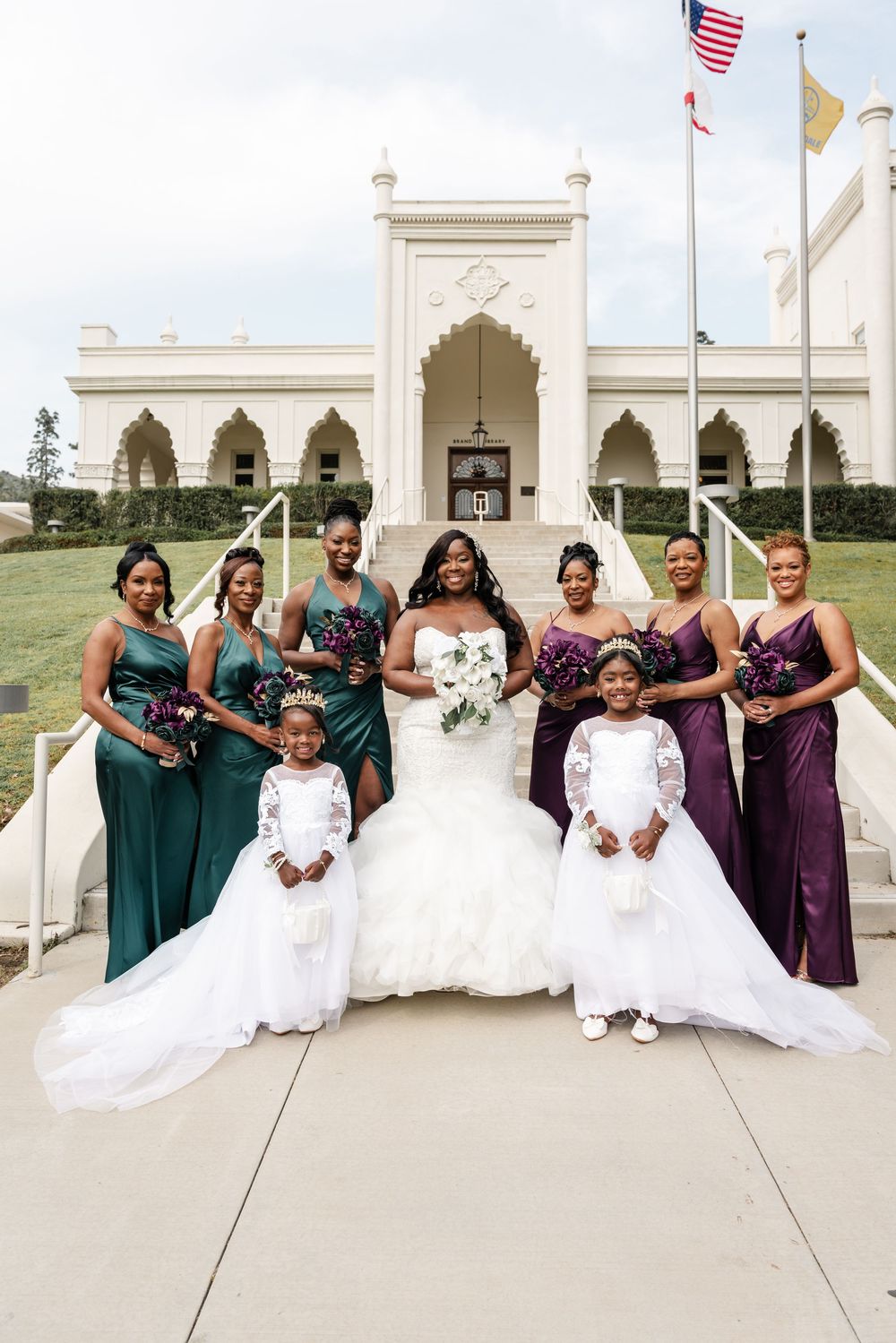 Wedding party poses outside ornate white building with American flag, featuring bridesmaids in green and purple dresses.