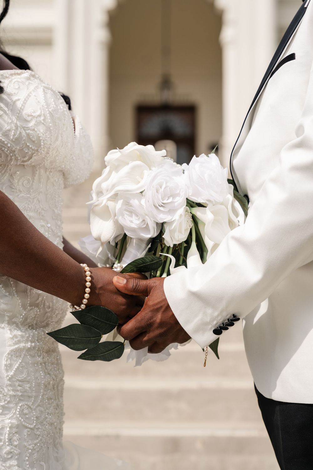 Close-up of couple's hands holding a white rose wedding bouquet, with bride in lace dress and groom in white jacket.