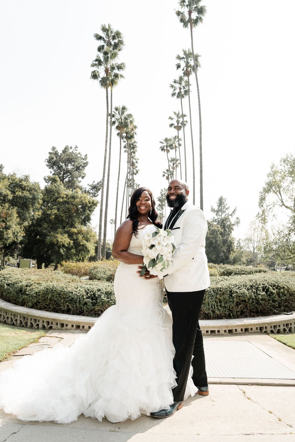 A happy couple shares an intimate moment in their wedding attire against tall palm trees and manicured gardens.