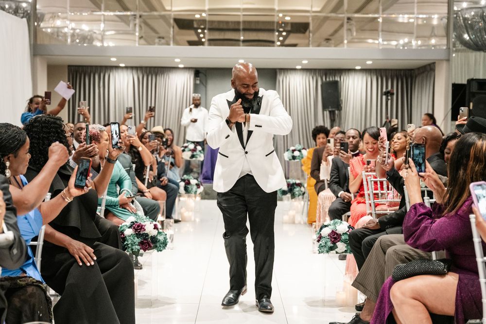 A model walks down a fashion show runway in a white dinner jacket as audience members take photos from their seats.
