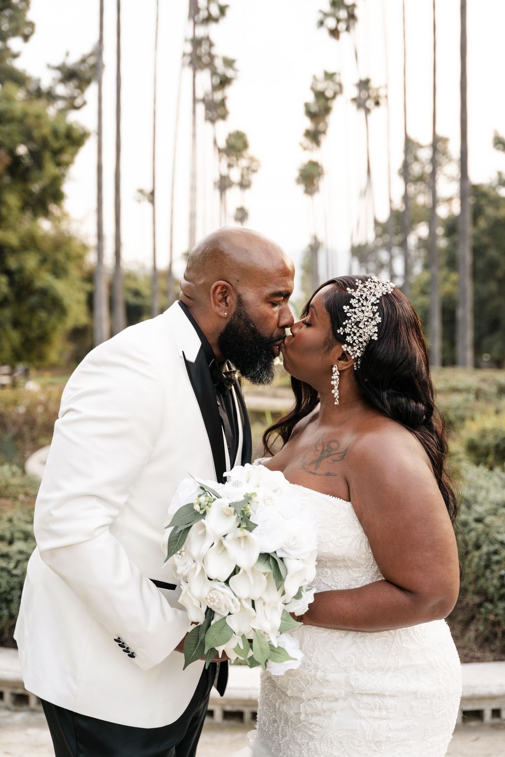 Happy newlyweds share a romantic kiss outdoors with a white floral bouquet in front of tall palm trees.