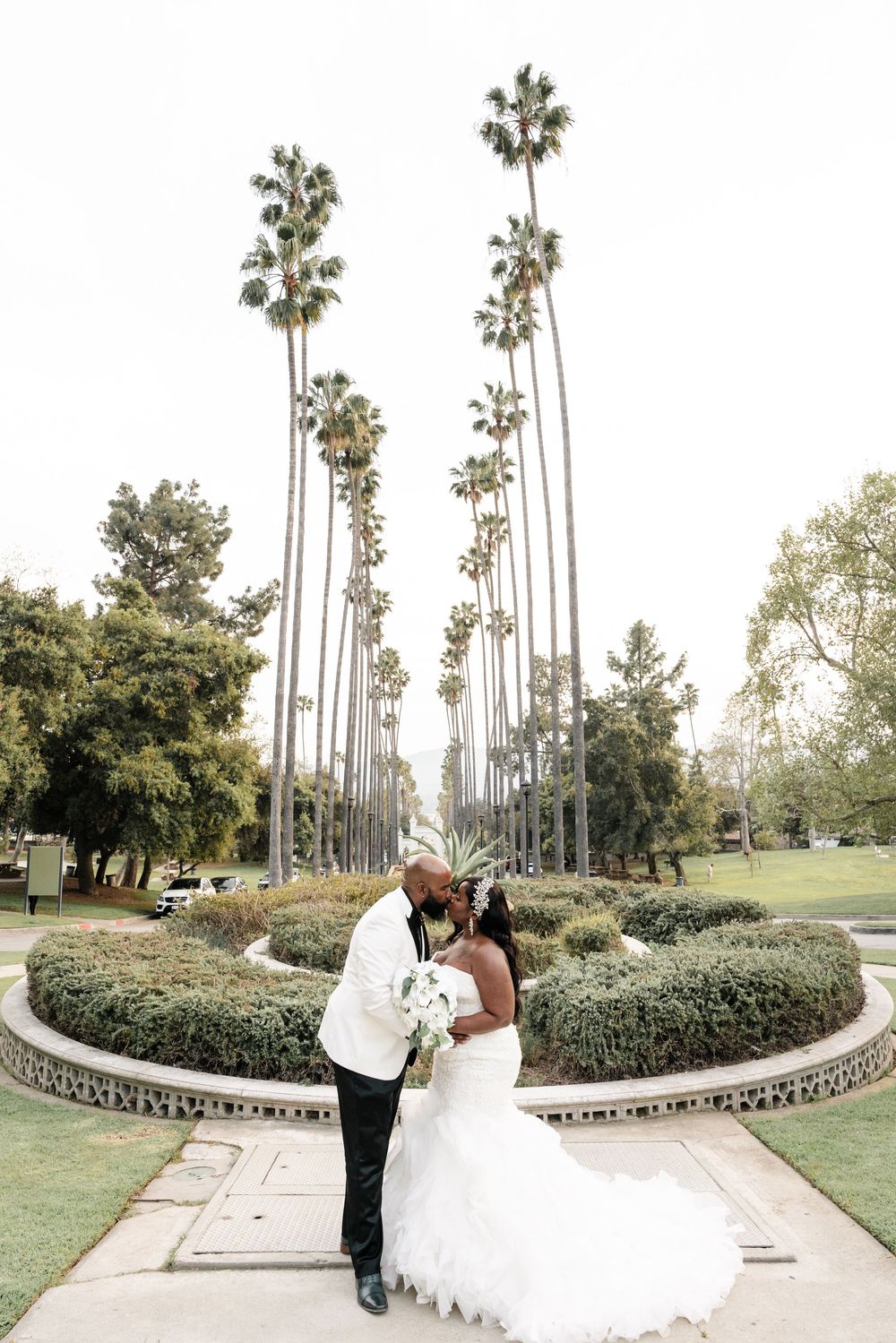 Couple shares a romantic moment near a stone planter surrounded by tall palm trees in a scenic garden setting.