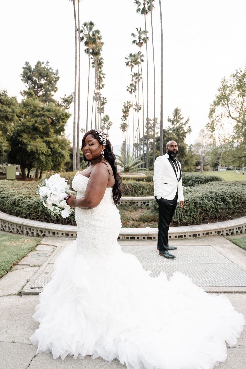 Two individuals pose for a romantic wedding photo in a garden setting framed by tall palm trees and lush landscaping.