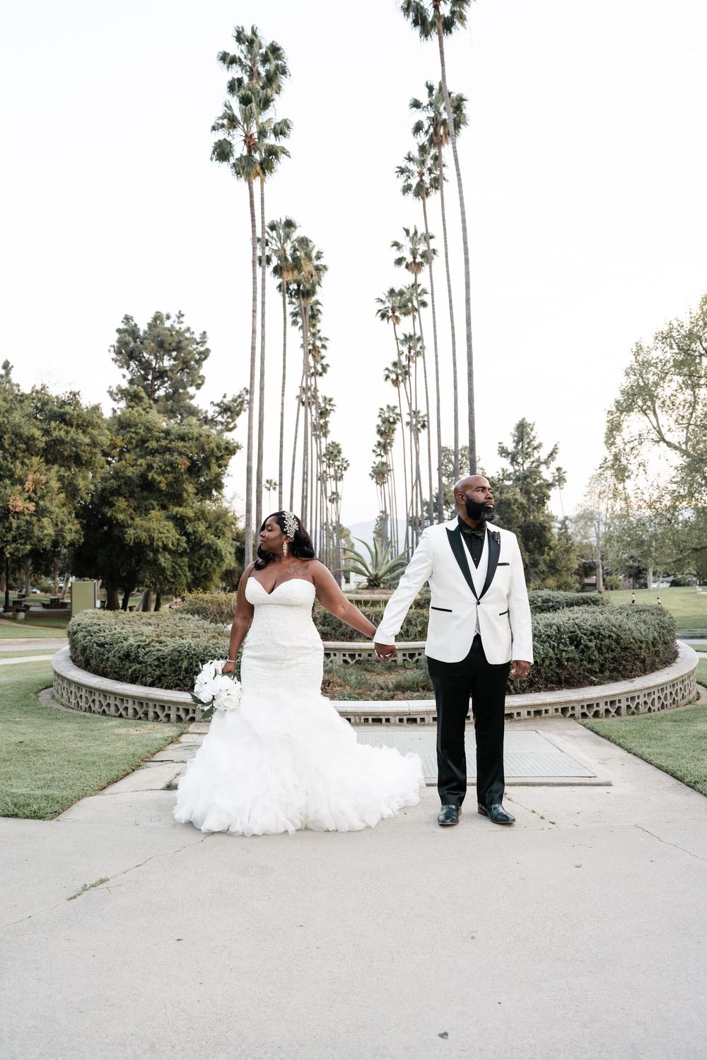 A couple stands together on a walkway surrounded by tall palm trees in an elegant outdoor wedding setting.